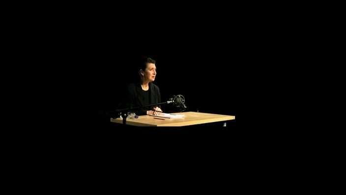 Still from The List play during Stellar Quines Rehearsal Rooms live stream by Inner Ear — a woman sits at a desk, starkly lit and alone in a black box theatre setting